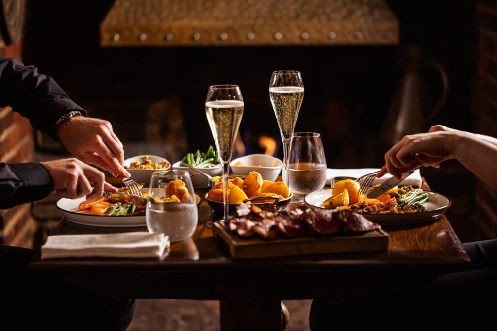 Two guests dining at a table with a festive roast, seasonal sides and glasses of Champagne at The Hind’s Head.