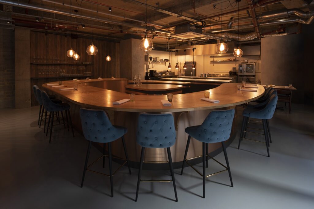 A curved wooden counter with upholstered stools faces an open kitchen at Behind, softly lit by hanging glass pendants.