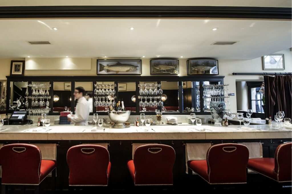 The marble counter at Bentley’s Oyster Bar & Grill, lined with red leather stools, hanging glassware and a bustling bar backdrop.