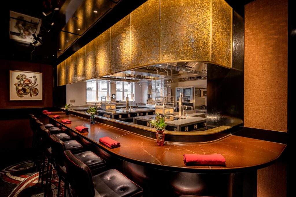 A curved leather counter facing an open kitchen beneath a gold-toned canopy at Cocochine, with red napkins set at each seat.