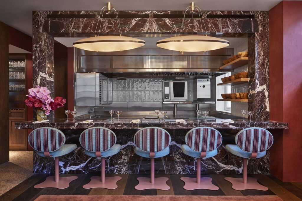 The chef’s counter at Muse by Tom Aikens, with marble surfaces, pastel bar stools and an open kitchen framed by warm lighting.