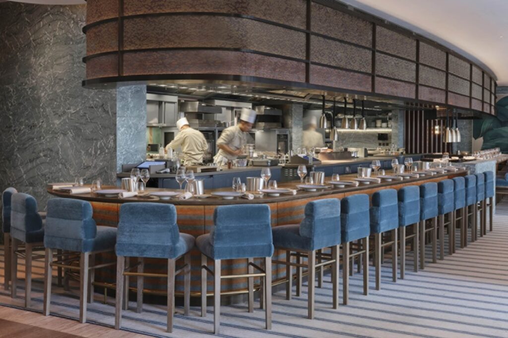 An open kitchen restaurant with chefs at work behind a curved counter lined with blue velvet stools and neatly set place settings.